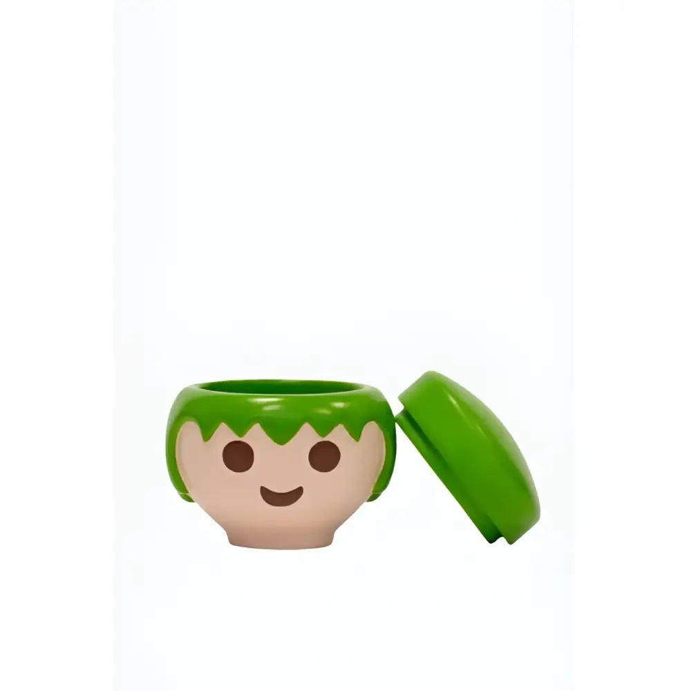 Tête Playmobil en plastique vert, sourire et cheveux courts, dans boîte verte au design ludique