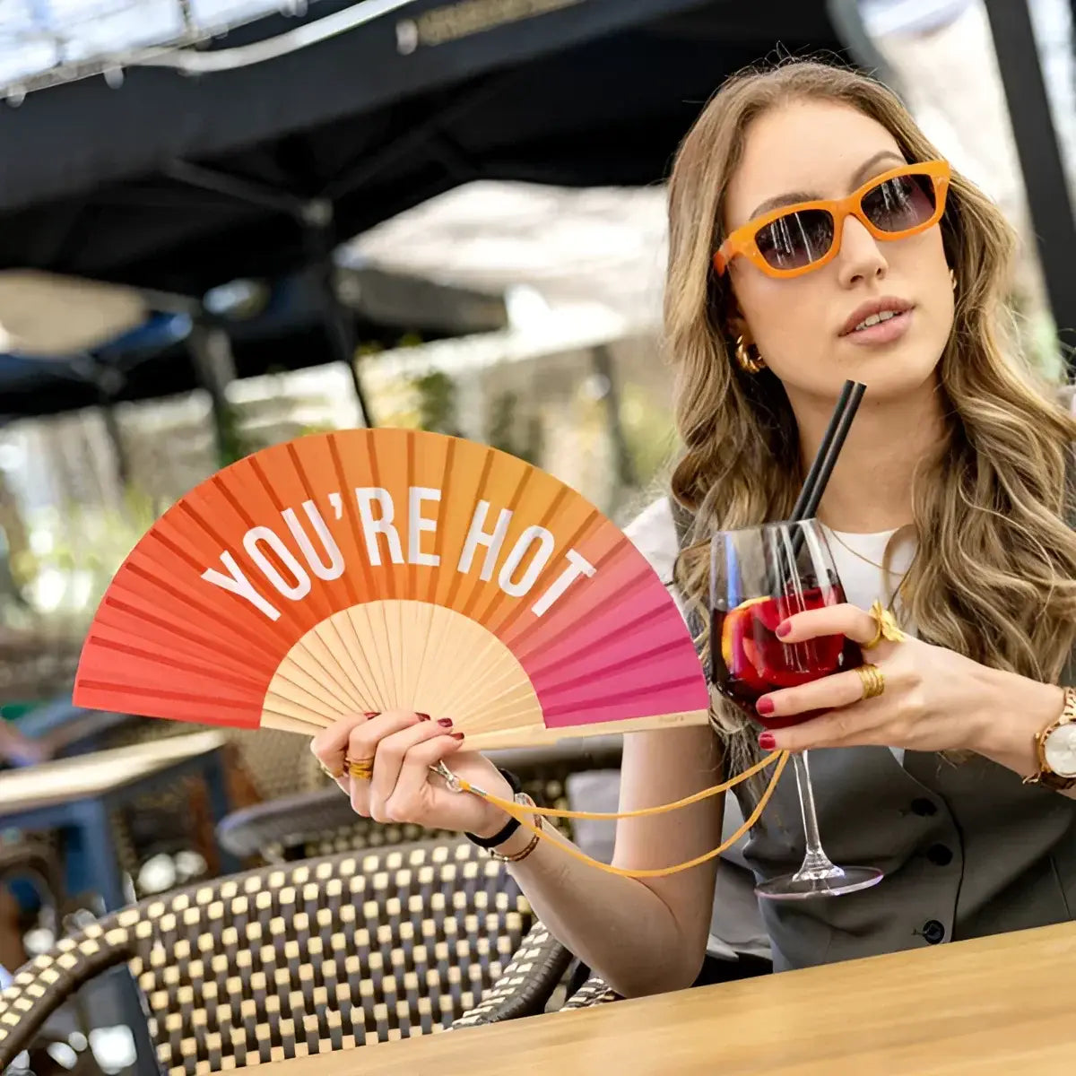 Femme en lunettes orange tenant éventail You’re Hot et sangria rouge