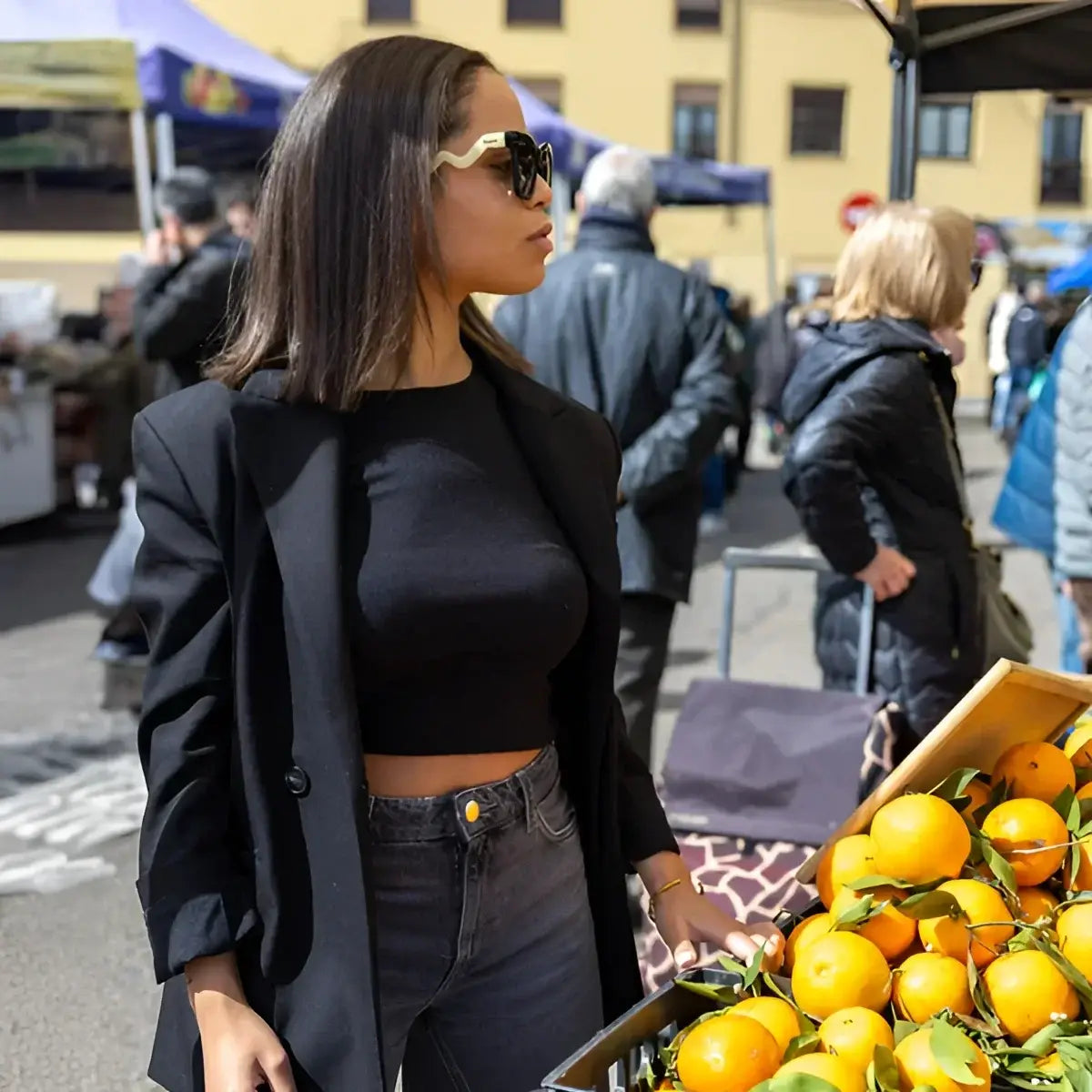 Femme en lunettes All You Need is Love, blazer noir, crop top, jeans sombres près de caisse d’oranges