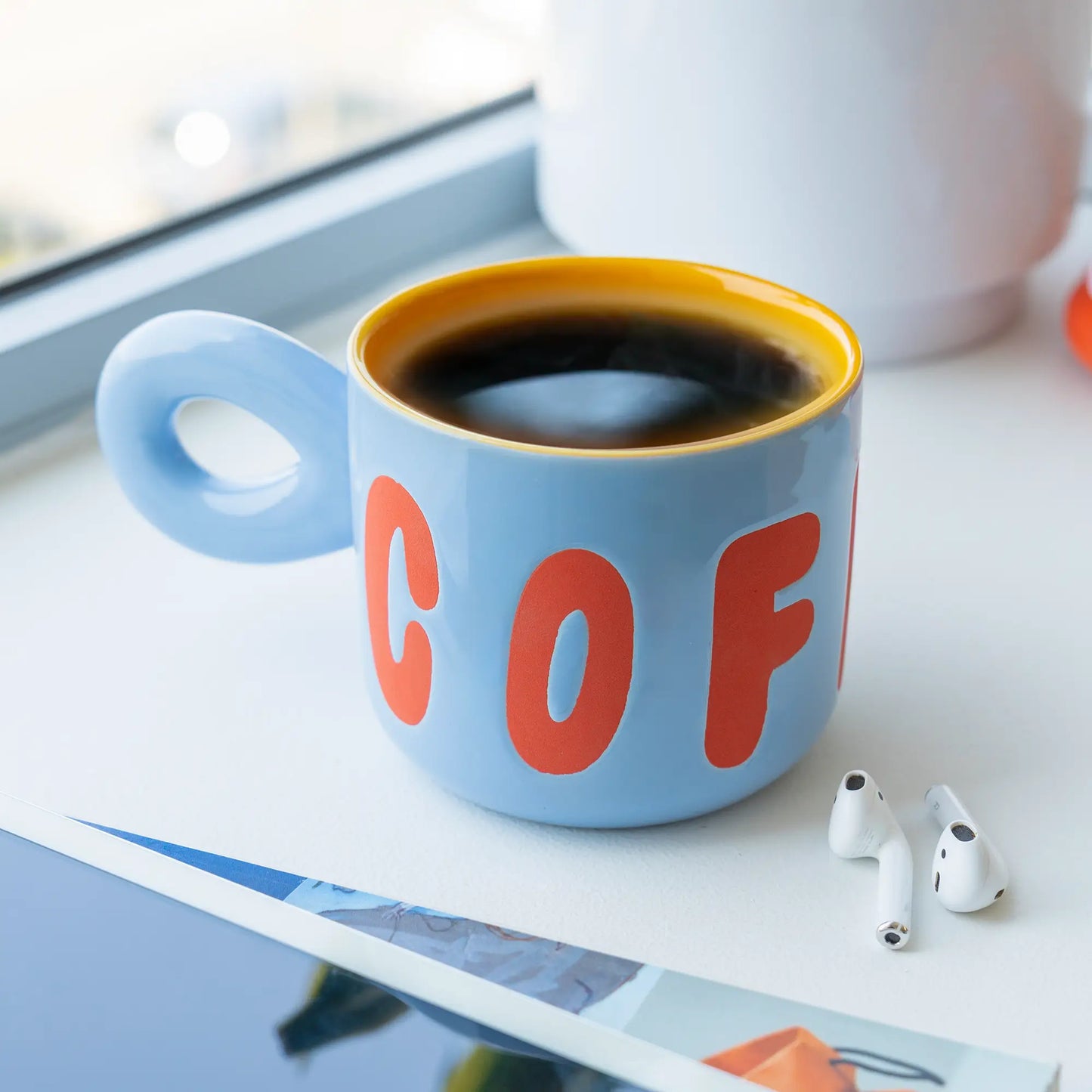 Tasse en céramique bleu clair, design épuré avec lettrage orange cof et intérieur jaune, remplie de café