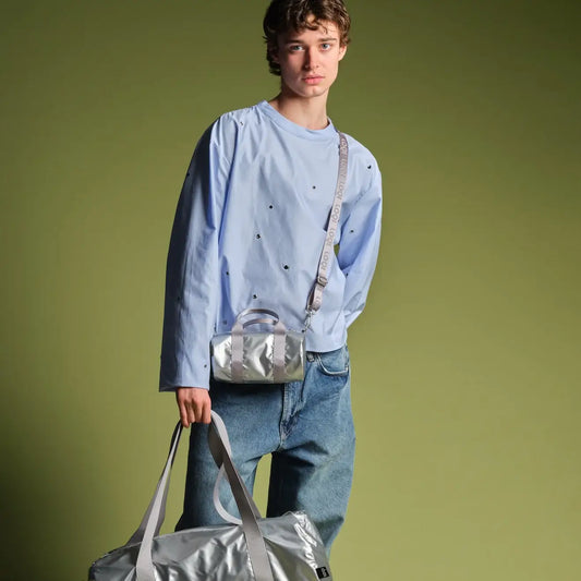 Jeune homme en chemise bleu clair à pois noirs, jeans oversized, sac week-end métallisé argenté élégant