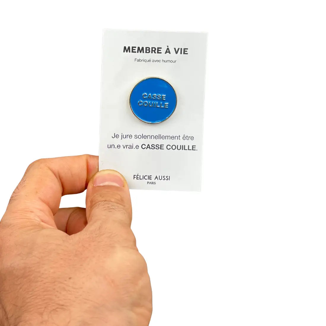 Main de personne tenant carte blanche avec pin bleu rond du produit Pin’s casse couille, touche d’humour