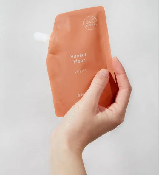 Pochette orange en plastique souple avec bec blanc, tenue à la main, recharge format pratique pour savon mains aux probiotiques Sunset Fleur